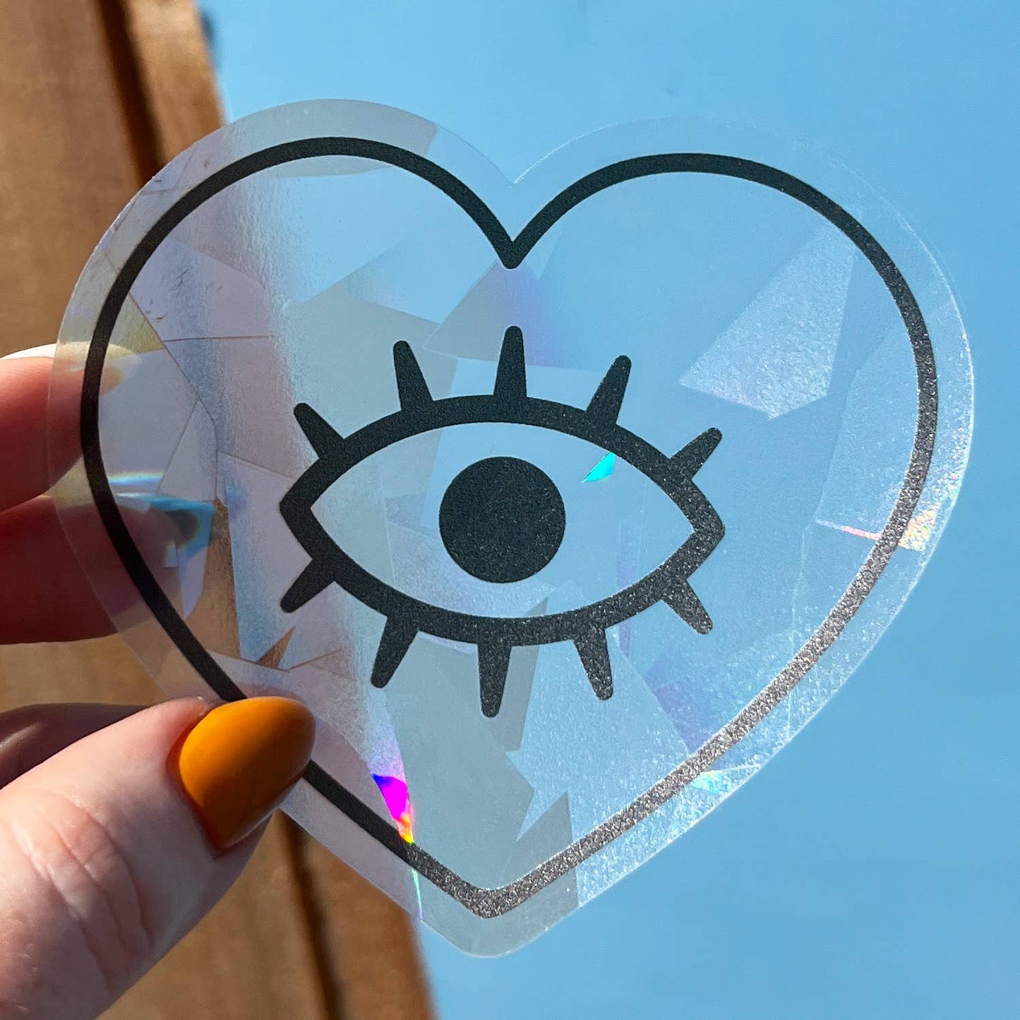 Heart-shaped suncatcher decal with an eye design held by a hand against a blue sky.
