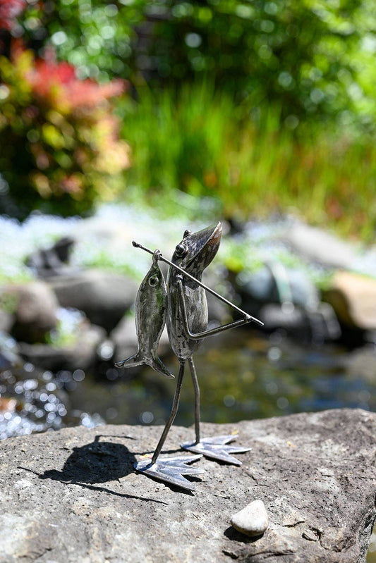 Metallic frog holding a fishing pole with a fish on the end standing on a rock by the river.