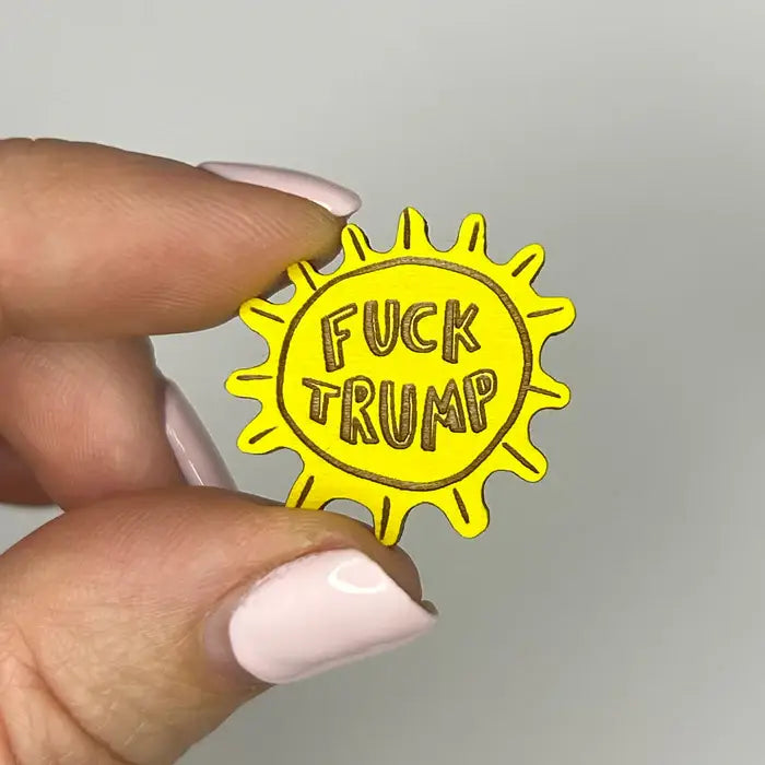 Yellow sun-shaped pin with 'FUCK TRUMP' text held by a hand