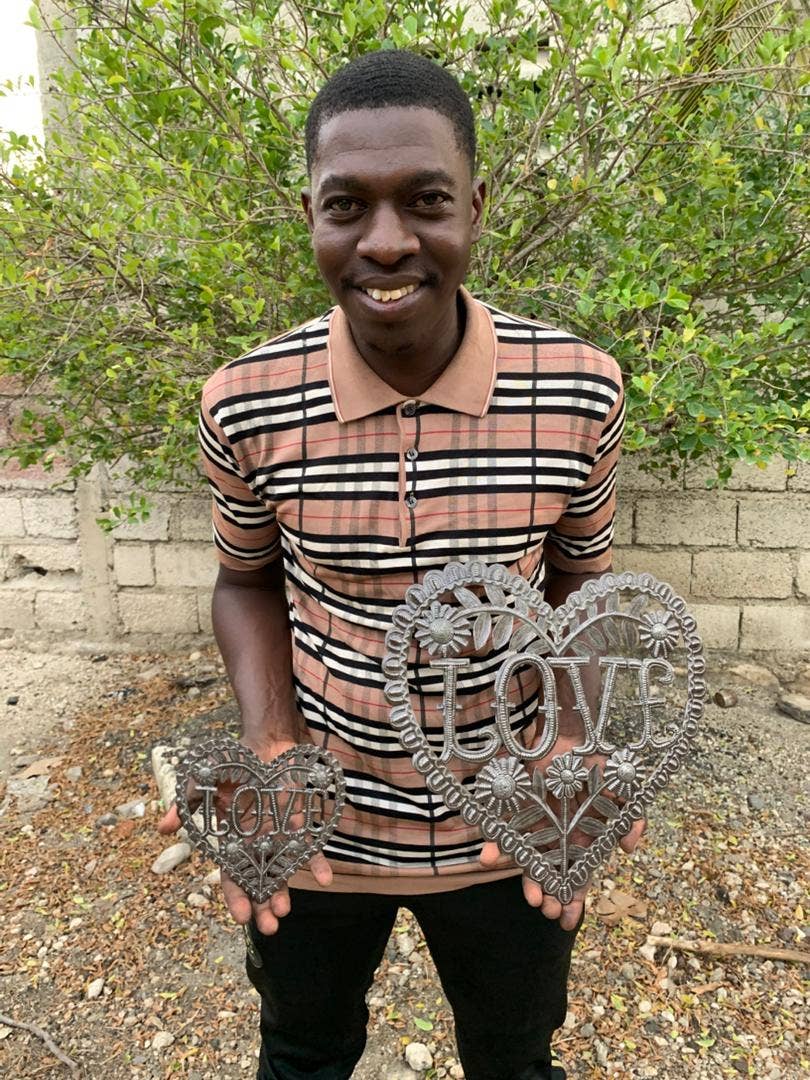 Artist holding 2 sizes of the handmade Haitian hammered metal wall art of a heart with the word Love surrounded by flowers