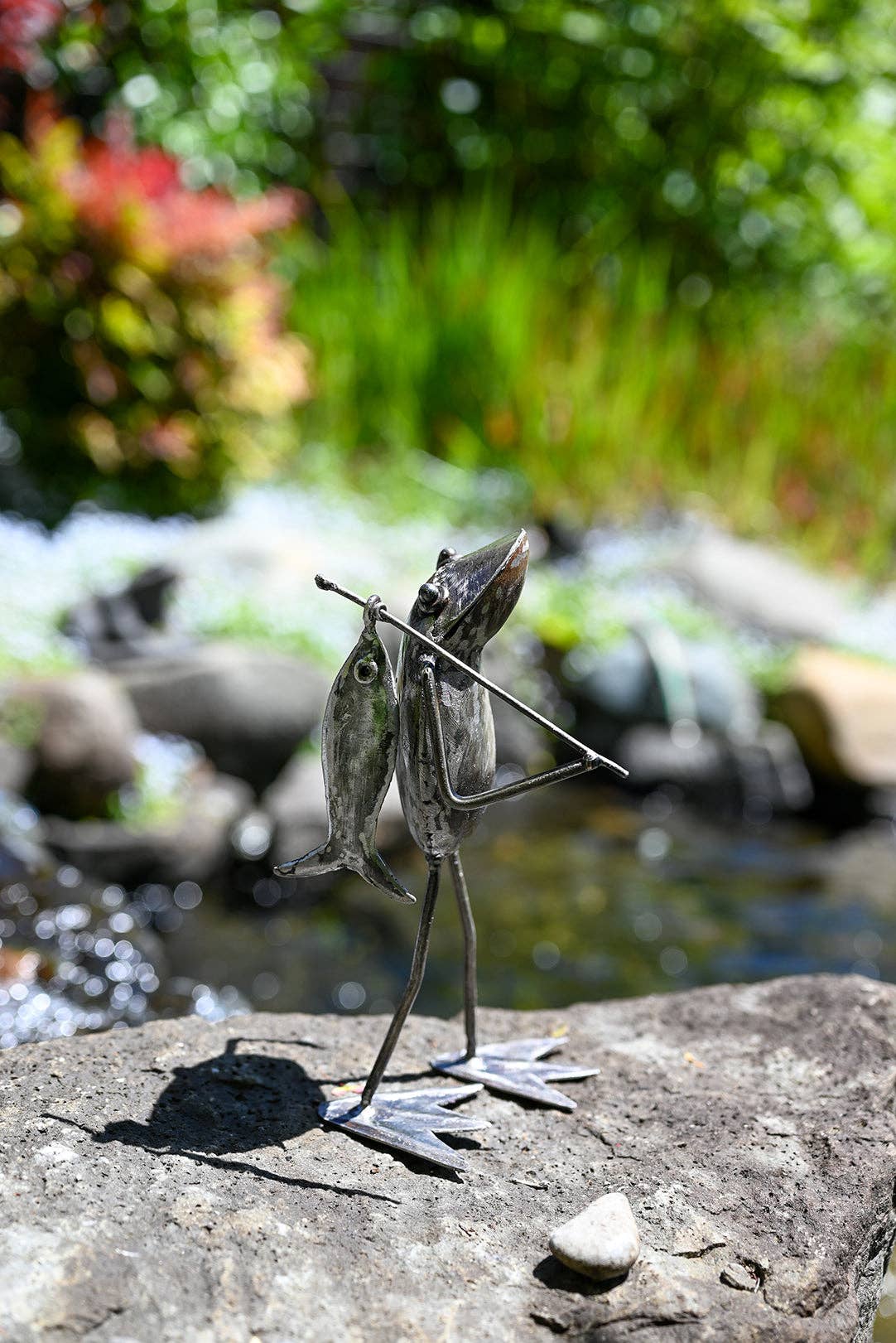 Metallic frog  holding a fishing pole with a fish on the end standing on a rock by the river.