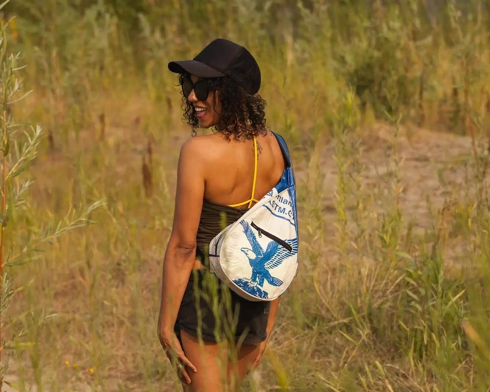 Person wearing a teardrop shaped sling bag. Exterior material is upcycled from feed and cement bags, Interior material is recycled from plastic bottles. Each design is unique.