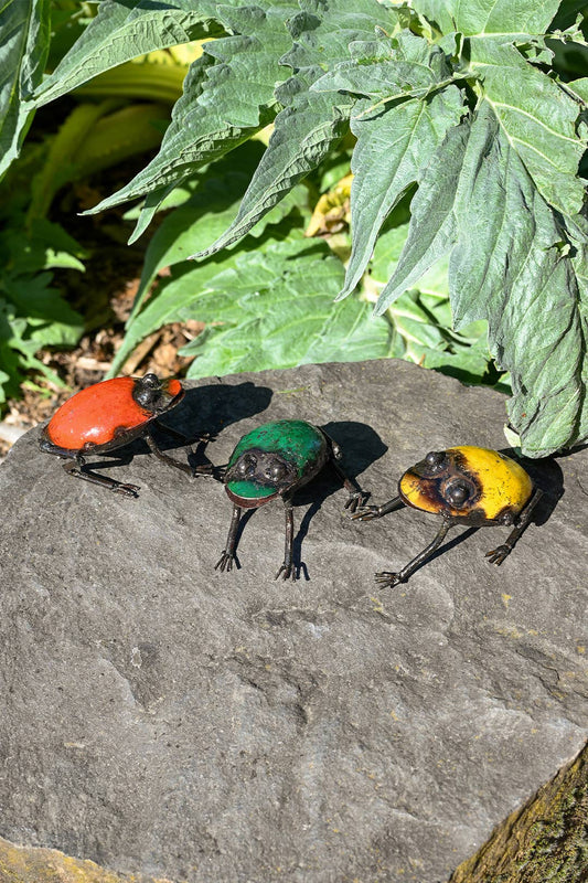 Three metallic frogs on a stone outside - red, green, and yellow.