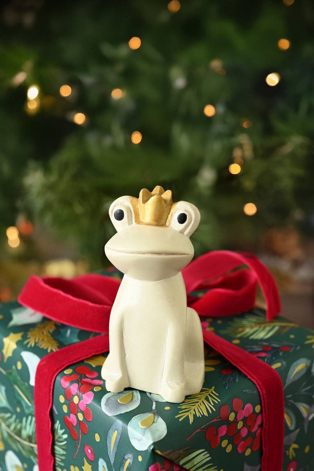 White Ceramic frog figurine with a gold crown sitting on a present in front of a Christmas tree.
