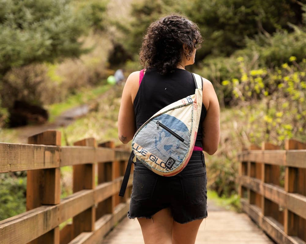 Person wearing a teardrop shaped sling bag. Exterior material is upcycled from feed and cement bags, Interior material is recycled from plastic bottles. Each design is unique.