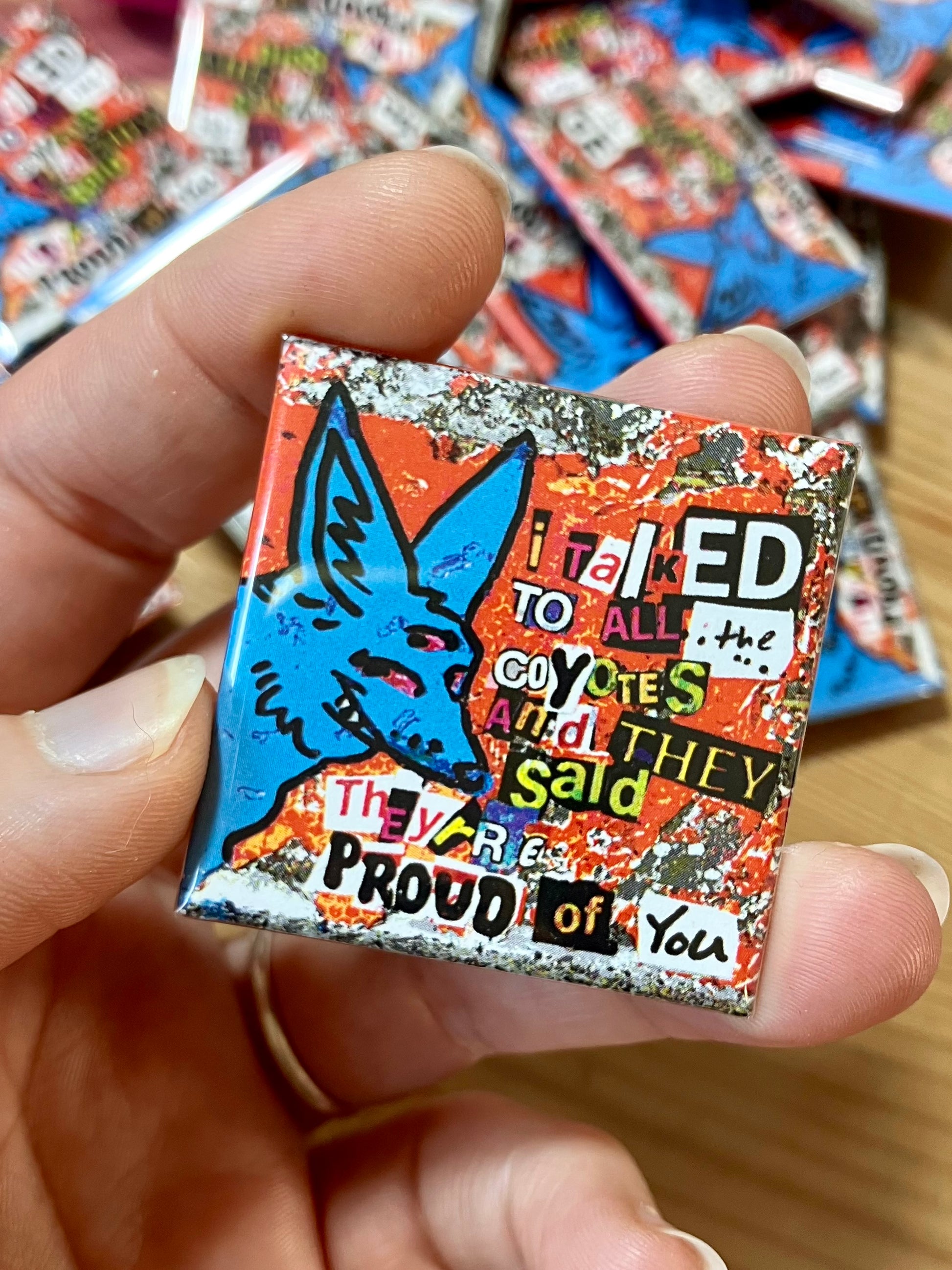 Hand holding a pin of a three eyed blue coyote that reads "I talked to all the coyotes and they said they're proud of you" using a ransom note-esk font