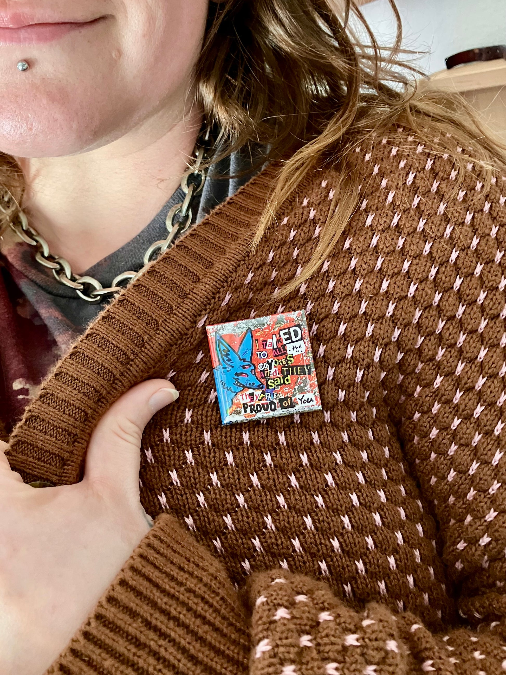 Person wearing a pin with a three eyed blue coyote that reads "I talked to all the coyotes and they said they're proud of you" using a ransom note-esk font