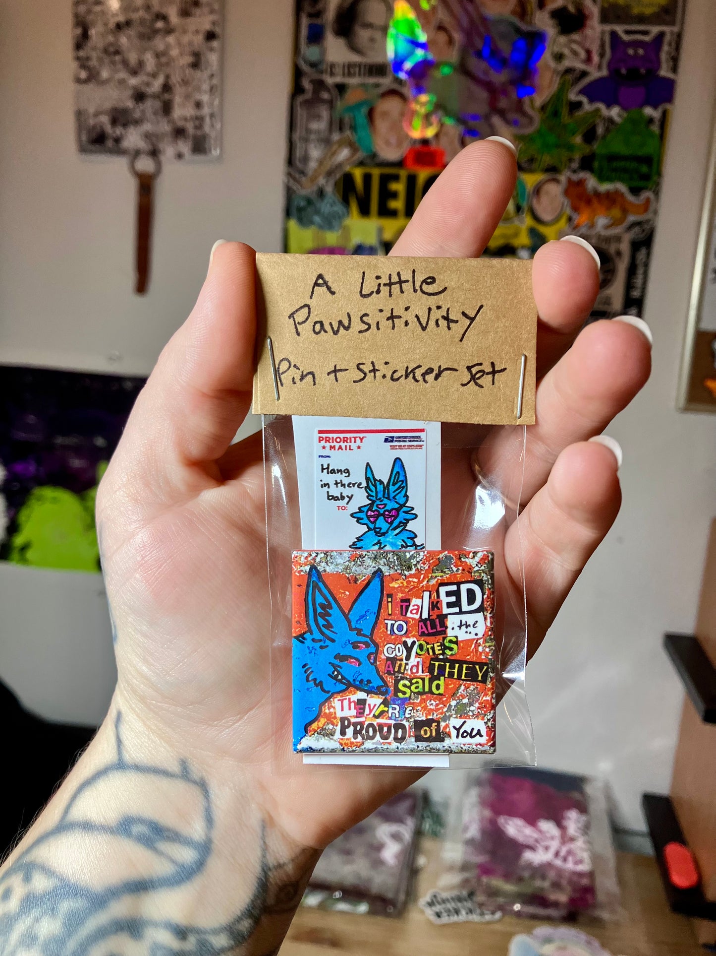 Hand holding a small package with a label reading 'A Little Pawsitivity Pin + Sticker Set'