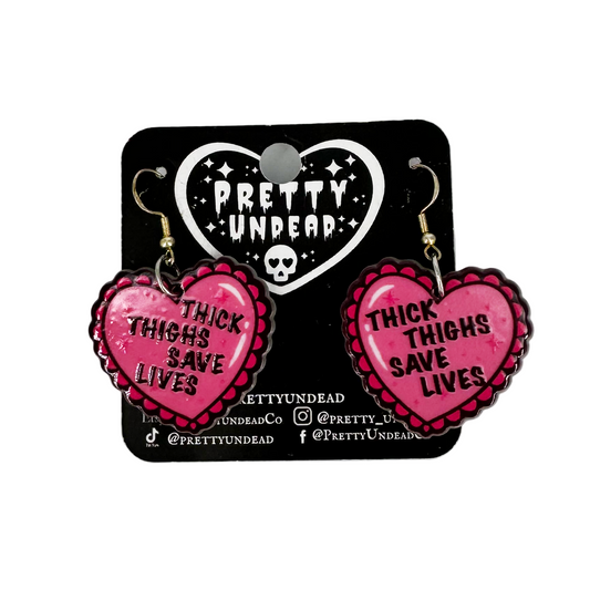 Pink heart-shaped earrings with 'Thick Thighs Save Lives' text on a Pretty Undead branded card.
