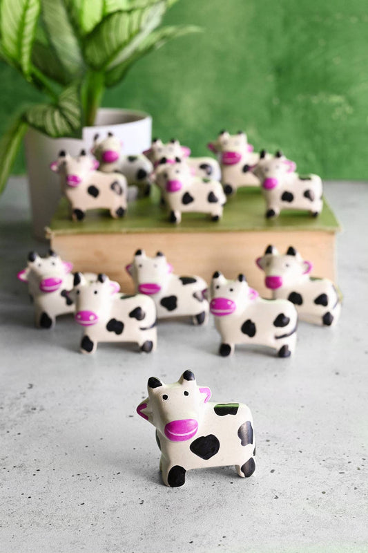 A dozen handcrafted soapstone cows with pink snouts and ears on a playful background made to look like field using a green book and a distant plant.
