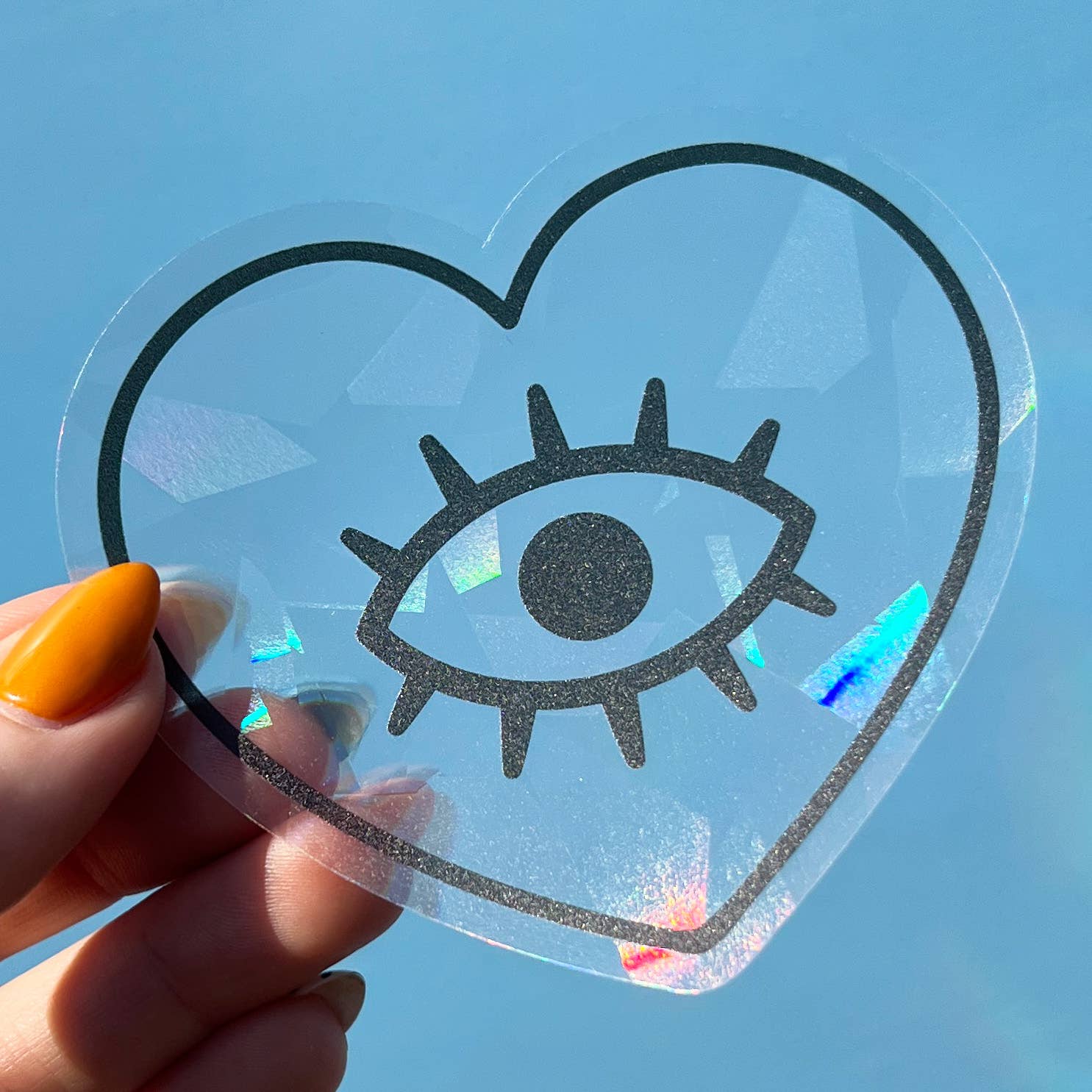Heart-shaped suncatcher decal with an eye design held by a hand against a blue sky.