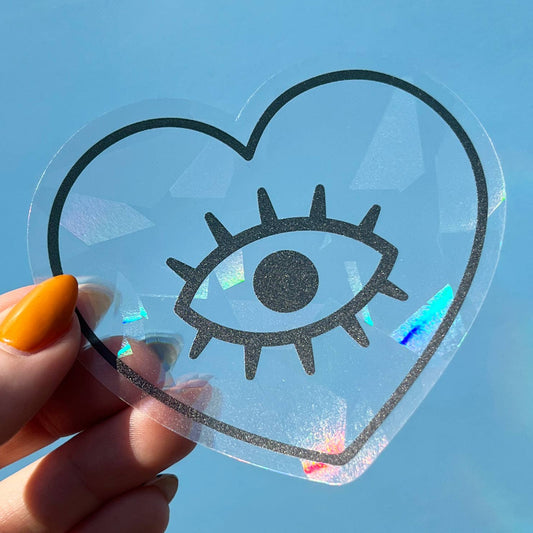 Heart-shaped suncatcher decal with an eye design held by a hand against a blue sky.