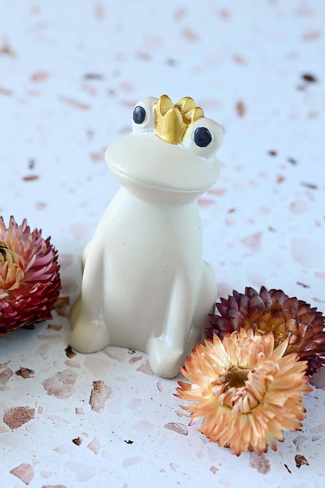 White Ceramic frog figurine with a gold crown sitting next to some flowers.