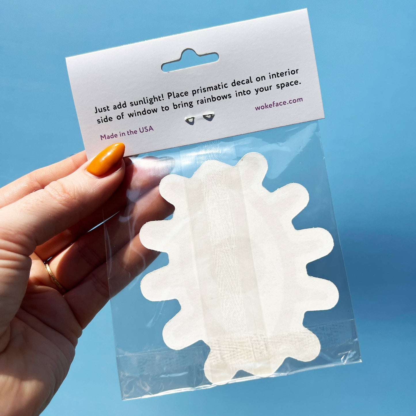 Hand holding the suncatcher decal inside it's package. Instructions read; Just add sunlight! Place prismatic decal on interior side of window to bring rainbows into your space. Made in the USA by wokeface.com