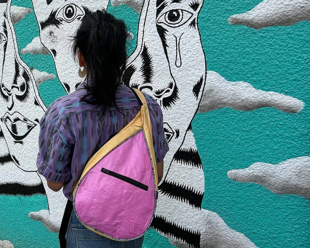 Person wearing a teardrop shaped sling bag. Exterior material is upcycled from feed and cement bags, Interior material is recycled from plastic bottles. Each design is unique.